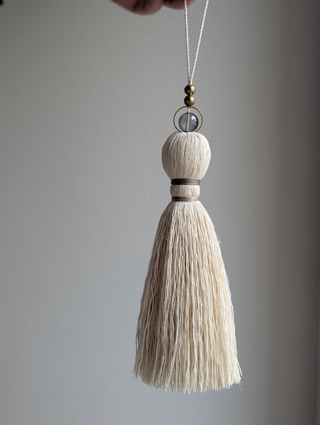 A close-up of a decorative hanging tassel with taupe colored thread, a gray glass bead and brass beads shown against a soft gray background.