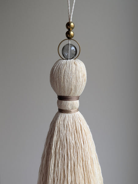 A close-up of a decorative hanging tassel with taupe colored thread, a gray glass bead and brass beads shown against a soft gray background.