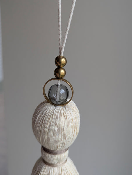 A close-up of a decorative hanging tassel with taupe colored thread, a gray glass bead and brass beads shown against a soft gray background.