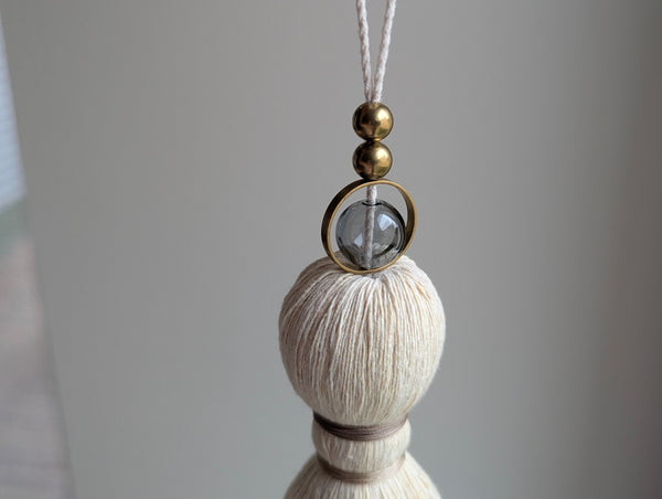 A close-up of a decorative hanging tassel with taupe colored thread, a gray glass bead and brass beads shown against a soft gray background.