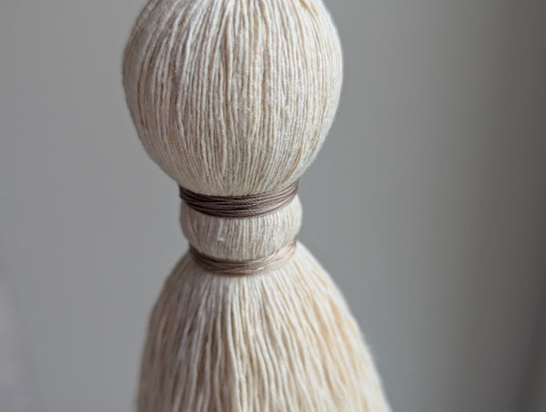 A close-up of a decorative hanging tassel with taupe colored thread, a gray glass bead and brass beads shown against a soft gray background.