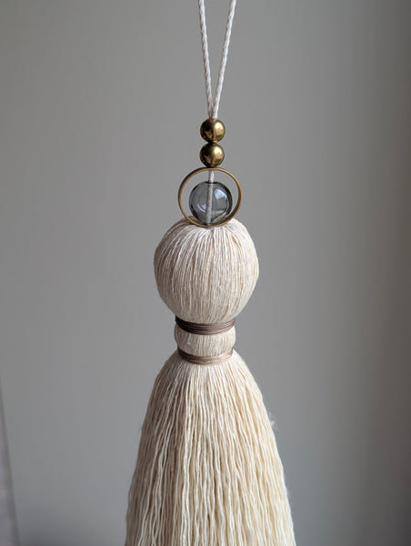 A close-up of a decorative hanging tassel with taupe colored thread, a gray glass bead and brass beads shown against a soft gray background.