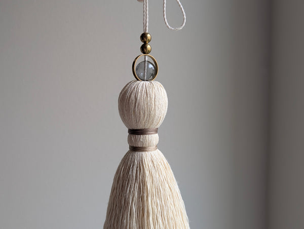 A close-up of a decorative hanging tassel with taupe colored thread, a gray glass bead and brass beads shown against a soft gray background.