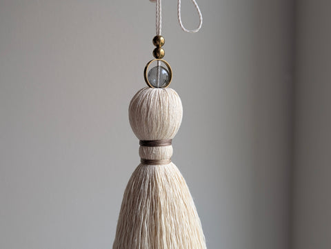 A close-up of a decorative hanging tassel with taupe colored thread, a gray glass bead and brass beads shown against a soft gray background.