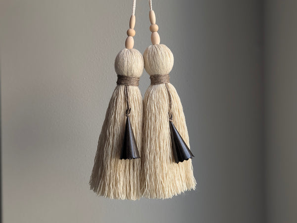 Two cream-colored cotton tassel tassels with natural wood beads and dark brown bells, hanging against a neutral gray background.