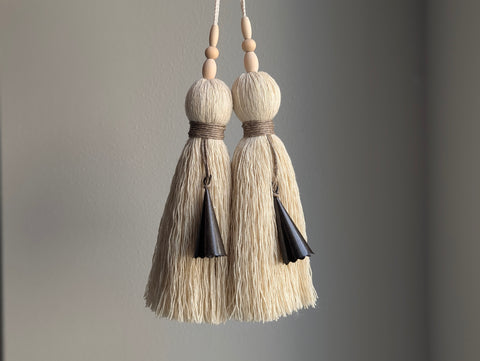 Two cream-colored cotton tassel tassels with natural wood beads and dark brown bells, hanging against a neutral gray background.