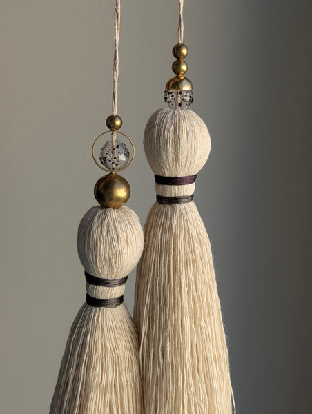 A close-up of a double tassel with deep purple colored thread, and glass and brass beads at the top against a neutral gray background