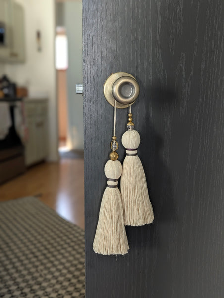 A close-up of a double tassel with deep purple colored thread, and glass and brass beads at the top against a dark door background