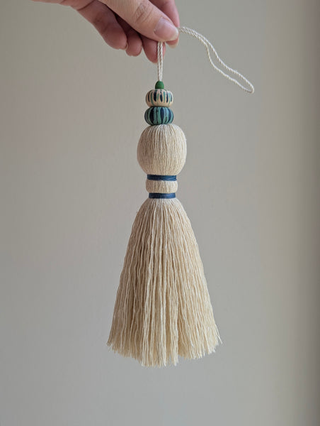 A close-up of a handmade wooden bead tassel, featuring a cream-colored cotton and blue and green accents against a soft beige background.