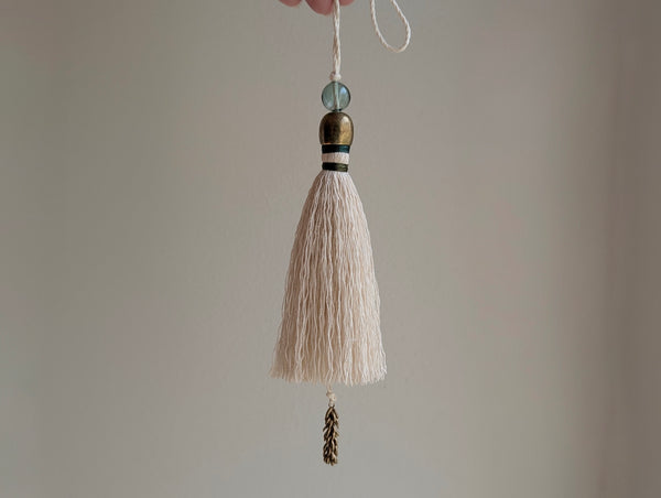 A cream-colored cotton tassel with green and brass accents held by a hand against a neutral beige background.