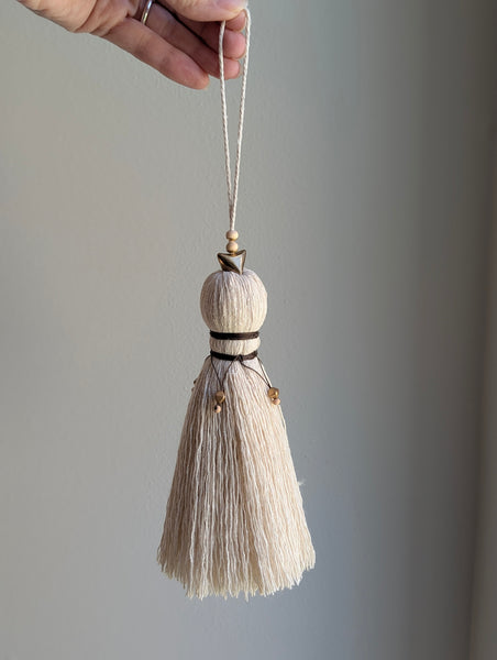A close-up of a decorative tassel with cream-colored thread, a brown wrapped detail, and brass and wooden beads shown against a neutral gray background