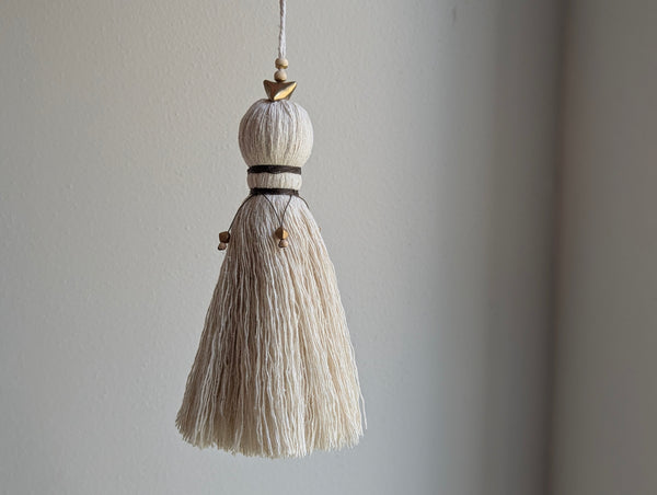 A close-up of a decorative tassel with cream-colored thread, a brown wrapped detail, and brass and wooden beads shown against a neutral gray background