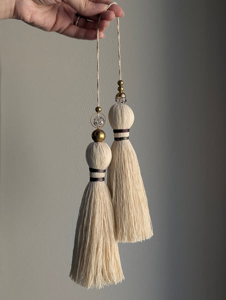 A close-up of a double tassel with deep purple colored thread, and glass and brass beads at the top against a neutral gray background