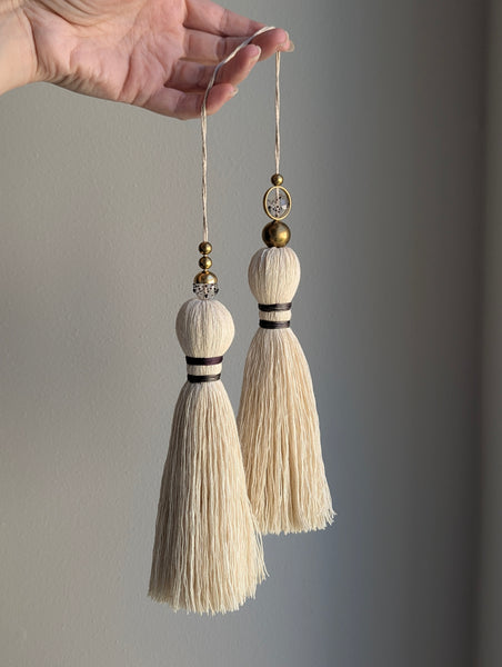 A close-up of a double tassel with deep purple colored thread, and glass and brass beads at the top against a neutral gray background