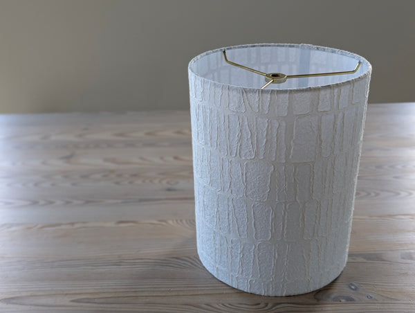 white paper textured lamp shade with rectangle pattern and brass hardware sitting on a light wood table