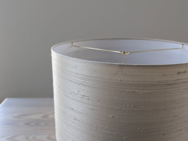 Close-up of a textured silk lampshade with brass hardware against a neutral background