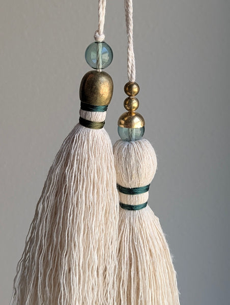 A decorative tassel featuring off-white cotton cord, two small metallic gold beads, one larger metallic gold bead, and a translucent green glass bead