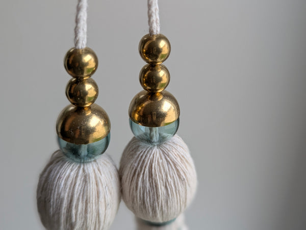 A decorative tassel featuring off-white cotton cord, two small metallic gold beads, one larger metallic gold bead, and a translucent green glass bead