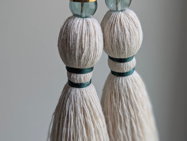A decorative tassel featuring off-white cotton cord, two small metallic gold beads, one larger metallic gold bead, and a translucent green glass bead