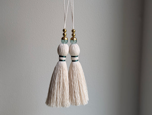 A decorative tassel featuring off-white cotton cord, two small metallic gold beads, one larger metallic gold bead, and a translucent green glass bead