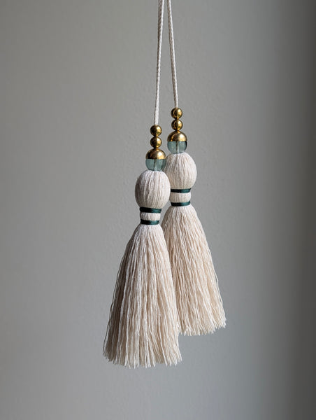 A decorative tassel featuring off-white cotton cord, two small metallic gold beads, one larger metallic gold bead, and a translucent green glass bead