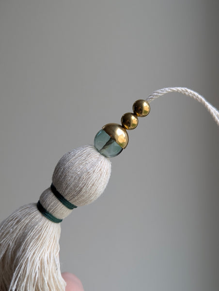 A decorative tassel featuring off-white cotton cord, two small metallic gold beads, one larger metallic gold bead, and a translucent green glass bead