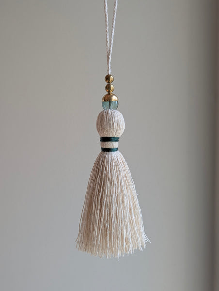 A decorative tassel featuring off-white cotton cord, two small metallic gold beads, one larger metallic gold bead, and a translucent green glass bead