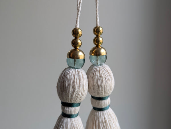 A decorative tassel featuring off-white cotton cord, two small metallic gold beads, one larger metallic gold bead, and a translucent green glass bead