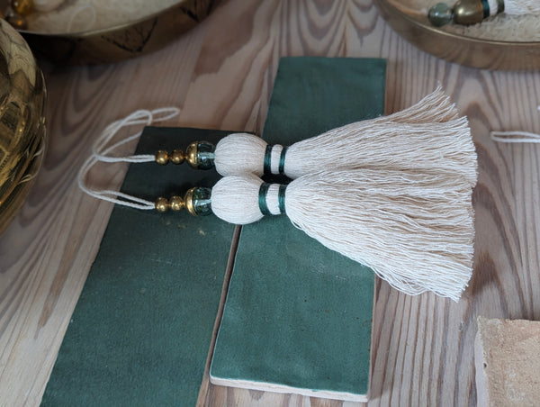 A decorative tassel featuring off-white cotton cord, two small metallic gold beads, one larger metallic gold bead, and a translucent green glass bead