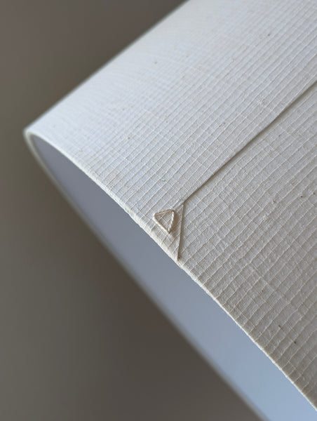 A close-up of a textured off-white fabric lamp shade with a small triangular stitched detail, shown against a soft neutral background.
