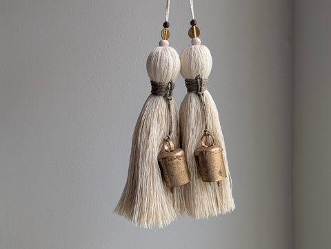 Two cream-colored tassel ornaments with small bronze bell charms hanging from cords against a plain gray wall.