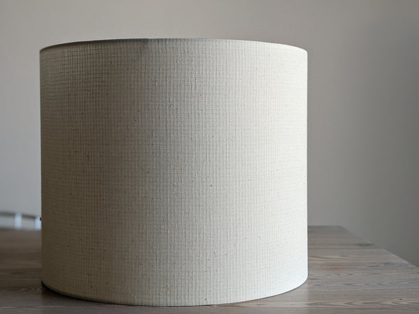 A large drum lampshade made of textured cream-colored fabric on a wooden table against a neutral gray background.