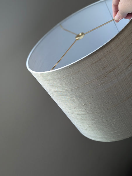 A close-up of a drum lampshade with a textured gray silk exterior and a white interior, held by a hand with brass spider washer hardware visible inside.