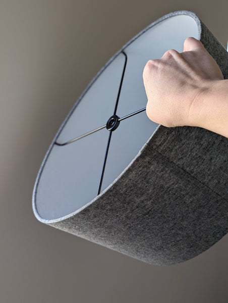 hand holding out an oval lamp shade in dark grey fabric with black hardware in a spider ring style meant to be used with a harp