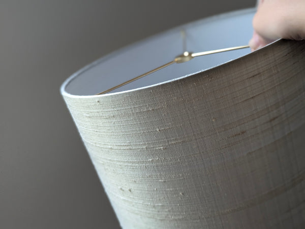 Close-up of a drum lampshade with a textured gray silk, showing a hand holding the top rim and a brass spider washer inside.