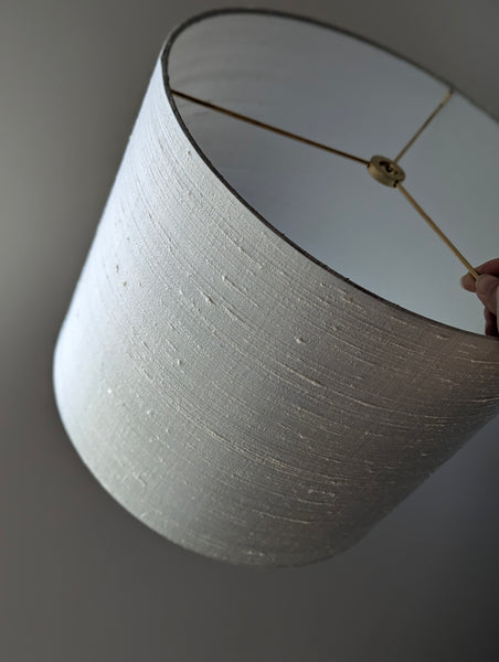 A close-up of a drum lampshade with a textured gray silk, shown against a neutral gray background. A hand is visible at the top right holding the shade.