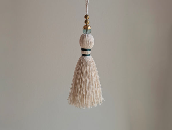 A decorative tassel featuring off-white cotton cord, two small metallic gold beads, one larger metallic gold bead, and a translucent green glass bead