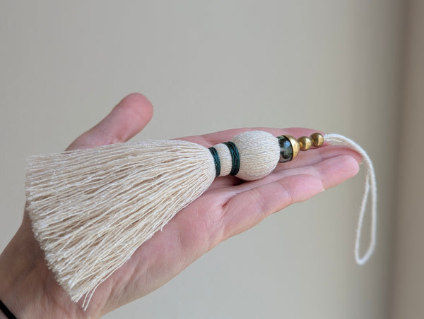 A decorative tassel featuring off-white cotton cord, two small metallic gold beads, one larger metallic gold bead, and a translucent green glass bead