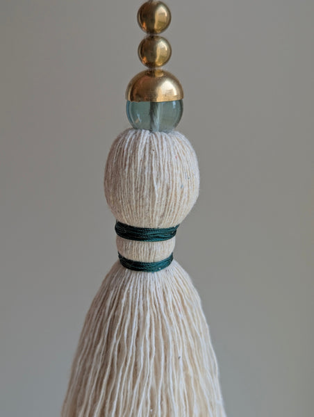 A decorative tassel featuring off-white cotton cord, two small metallic gold beads, one larger metallic gold bead, and a translucent green glass bead