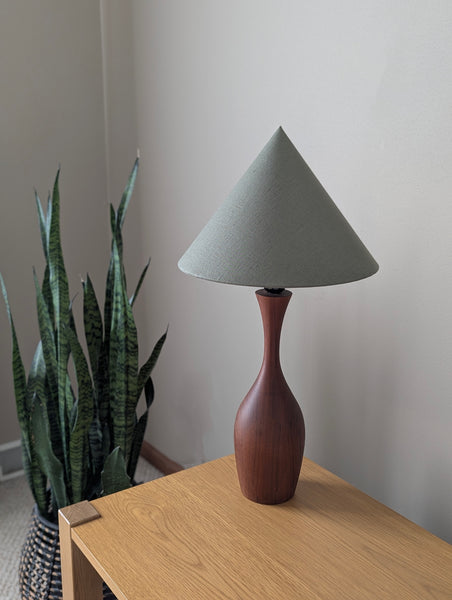 vintage walnut table lamp with a khaki green, cone-shaped lamp shade on a wooden nightstand next to a plant