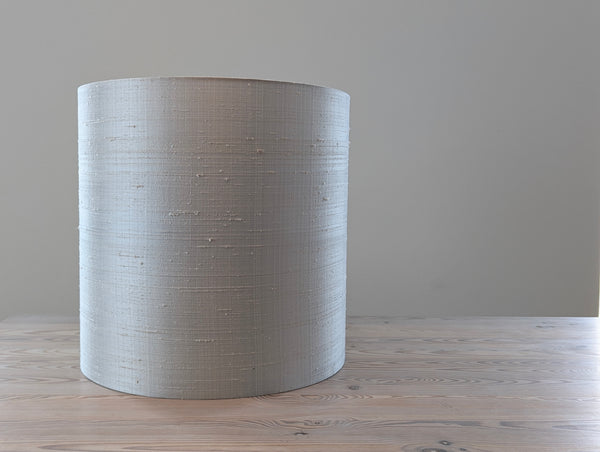 A tall, cylindrical drum lampshade made of gray silk, standing on a light wood surface against a neutral gray wall.