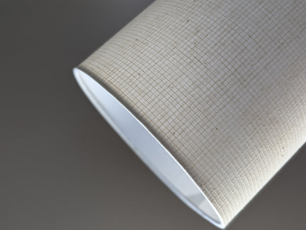 A close-up of a drum lampshade made of textured beige fabric with a fine grid pattern and a white inner lining, shown against a dark gray background.