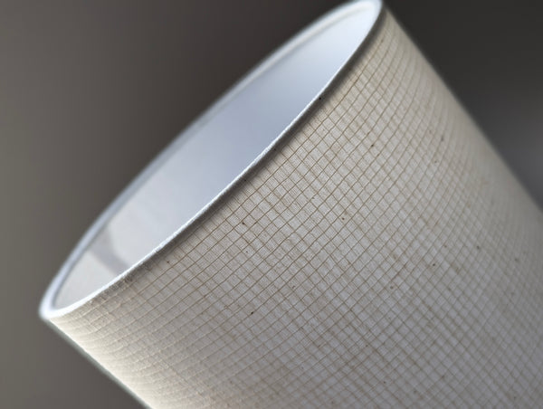 Close-up of a drum lampshade with a textured off-white fabric exterior and a smooth white interior, shown against a dark gray background.