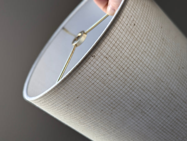 Close-up of a drum lampshade with a textured beige fabric exterior and a white interior, held by a hand near the rim.
