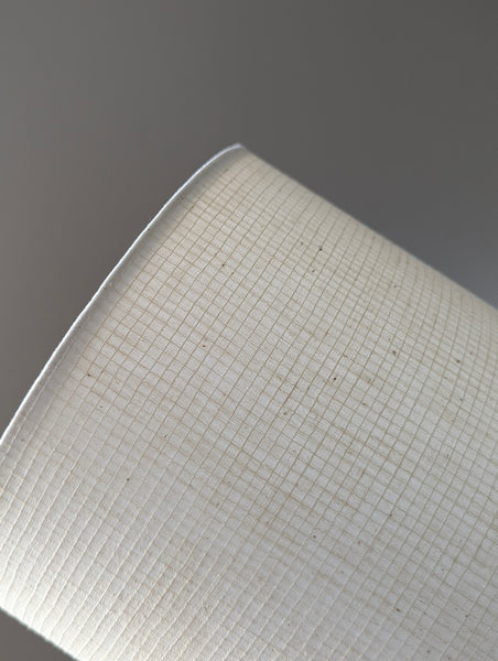 A close-up of a white textured lamp shade with a fine grid pattern, shown against a solid gray background.