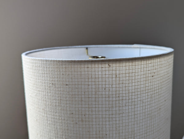 A close-up of a drum table lamp shade made of textured off-white fabric with a subtle grid pattern, showing a brass metal washer at the top against a neutral gray background.