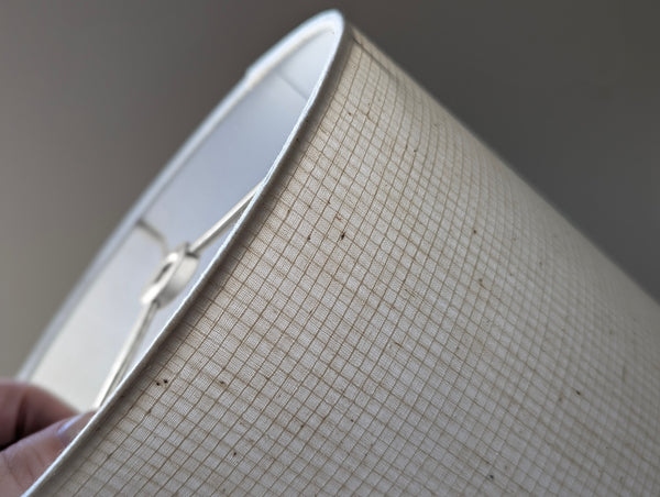 Close-up of a drum lampshade made of a beige woven or textured fabric with a fine grid-like pattern, held by a hand at the lower left edge.