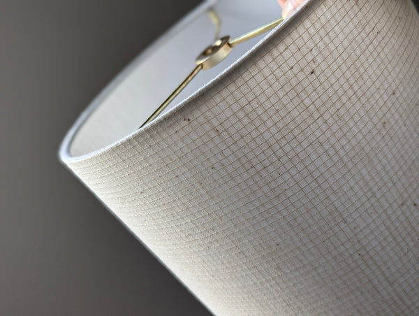 Close-up of a drum lampshade made of textured off-white fabric with a fine grid pattern, showing a gold-toned metal finial and part of the interior against a dark gray background.