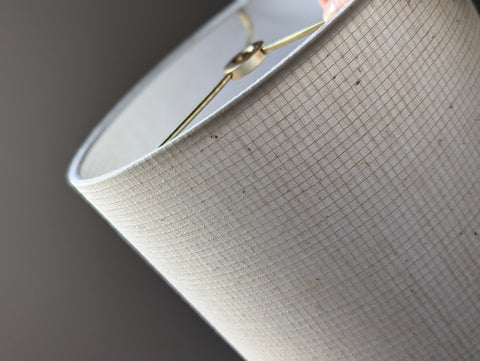 Close-up of a drum lampshade made of textured off-white fabric with a fine grid pattern, showing a gold-toned metal finial and part of the interior against a dark gray background.