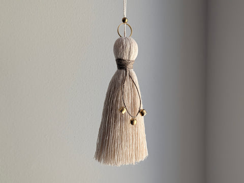 A close-up of a decorative tassel with cream-colored thread, a tan twine detail, and brass and wooden beads shown against a neutral gray background